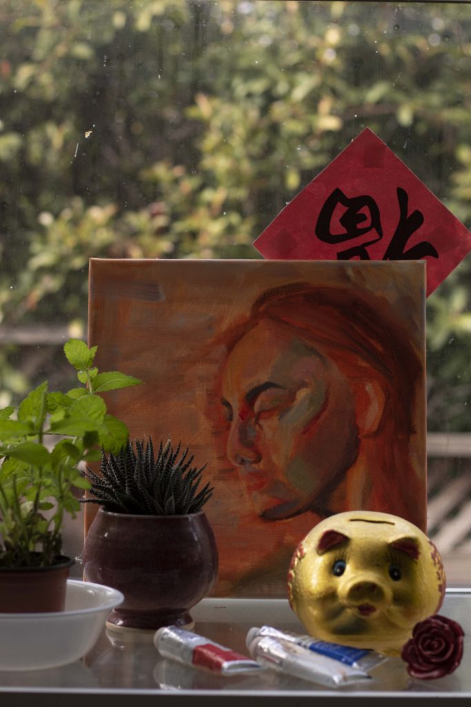 Photo of my culture, depicting a unfinished self-portrait with warm, color-blocked hues. Plants, oil paint tubes, a gold piggy bank, and a ceramic rose frame the background. A Chinese "fu" and bushes make up the background.