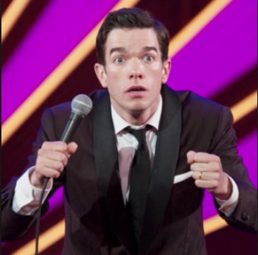Picture of John Mulaney on stage, a microphone in his right hand. Both of his hands are up in a motion to simulate driving. 