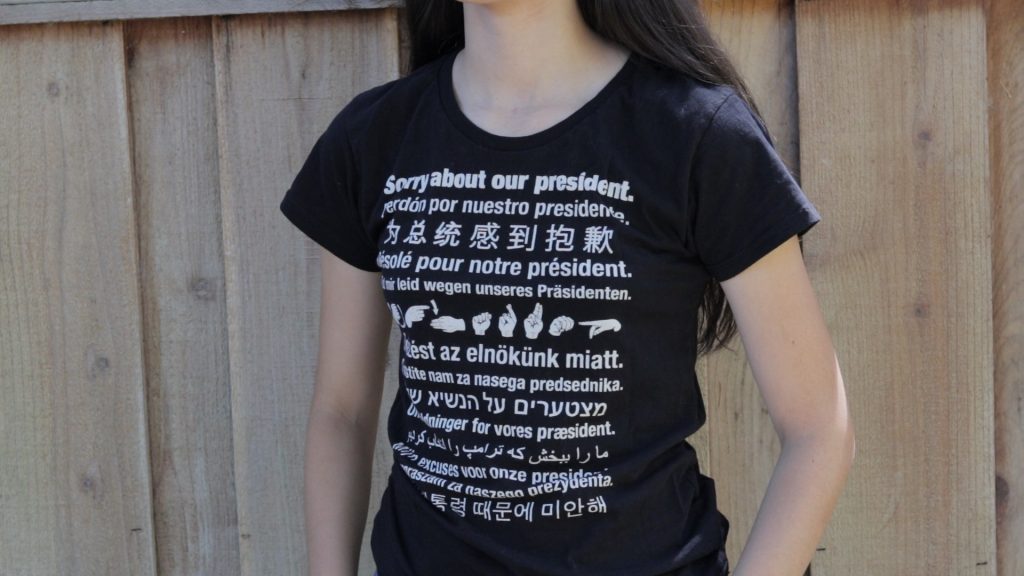 A girl wearing a tshirt that says Sorry about our president in a dozen different languages.