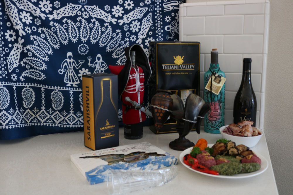 Image of Georgian food on a plate, food in a small bowl, a glass horn, a box of chocolates, some drinking horns, multiple bottles of wine, and a textile table cover in the background.
