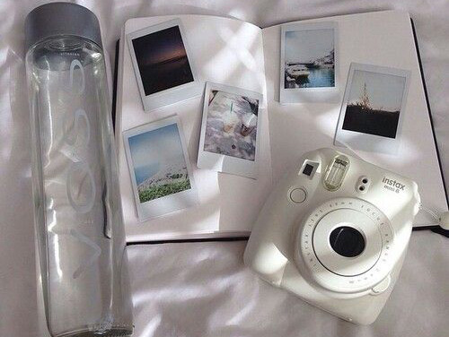 A white polaroid camera, a journal with polaroids inside, and a voss water bottle.