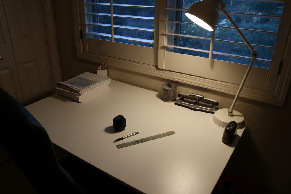 A photo showing warm light on a desk, directed onto a couple of items. a camera lens, ruler, and a pen. Showing that the mood is very chill, relaxing. Just a casual homework Sunday.