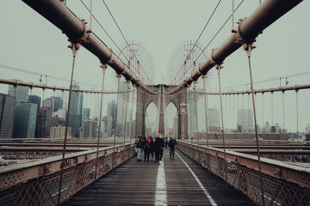 A photo of the Brooklyn Bridge. The perspective from the walkway of the bridge. Showing the leading lines from the side and the middle running down to the center of the screen.