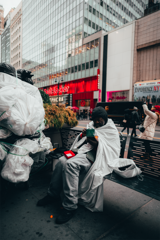 A photo of a homeless man sitting on a bench in the streets of New York, he has a cart full of garbage bags next to him.