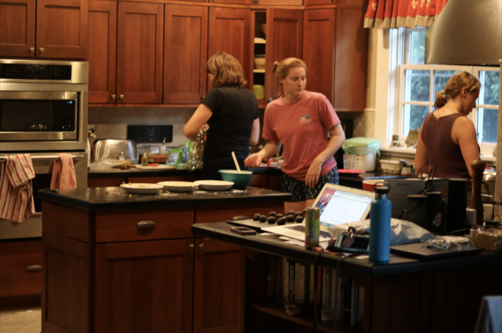 A photo of three women moving about a very messy kitchen, with plates, bowls, cups, a computer, and many other items strewn about.